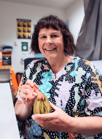 The image is of physiotherapist Rachel Andrews in a clinic room. She is wearing a colourful top and is holding a model of the female genitals.